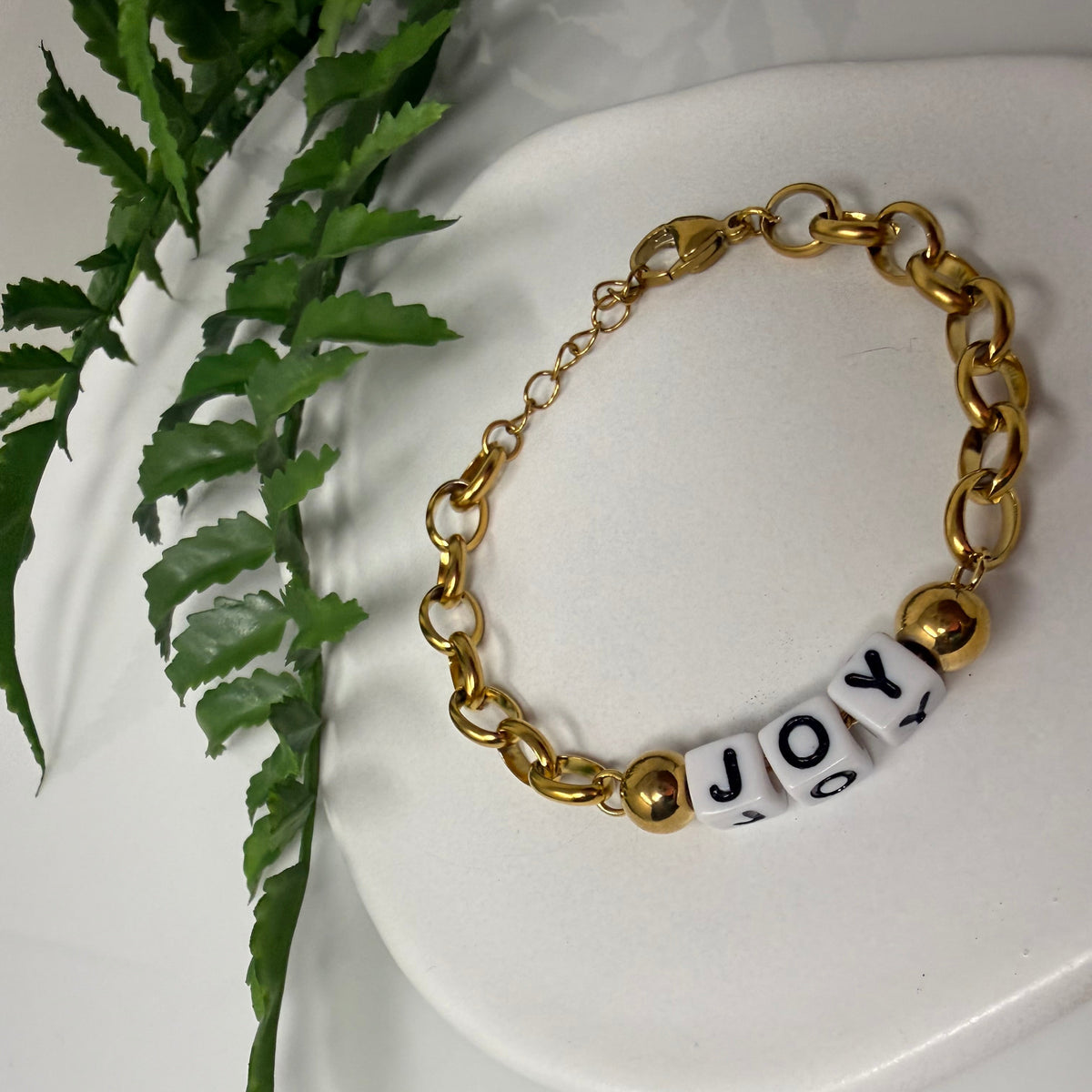 A gold-tone chain bracelet with three white cylindrical beads printed with the letters 'J', 'O', and 'Y', displayed on a white ceramic dish with green fern leaves in the background.